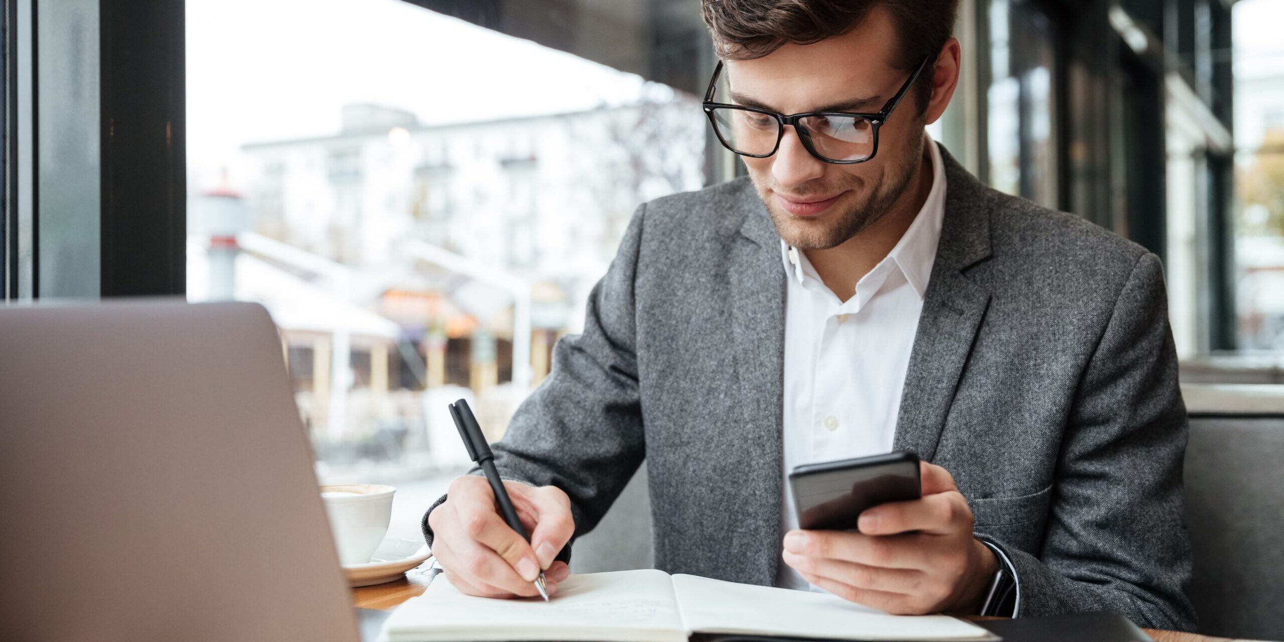 smiling-businessman-in-eyeglasses-sitting-by-the-table-in-cafe-with-laptop-computer-while-using-smartphone-and-writing-something-min