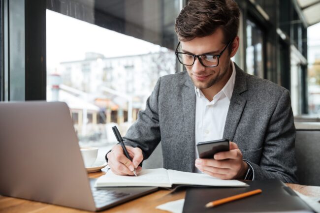 smiling-businessman-in-eyeglasses-sitting-by-the-table-in-cafe-with-laptop-computer-while-using-smartphone-and-writing-something-min